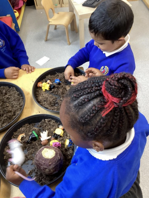 Two Nursery pupils, a boy and a girl, are pictured playing with some toy figures inside a bowl of soil, pretending that it is a farm.