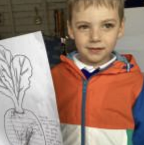 A male Nursery pupil is pictured smiling for the camera, whilst holding up a drawing of a Turnip he has drawn.