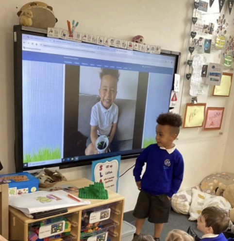 A young boy from Nursery is pictured standing at the front of his class, beside the Interactive Board on the wall, showing a photo of himself holding a plant.