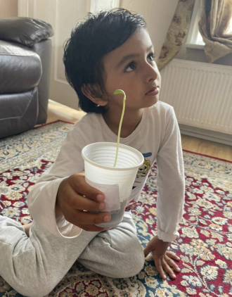 A boy from Nursery can be seen holding a plant in a pot, whilst sitting on the floor in his home.