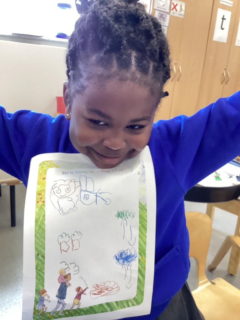 In this image, a young boy is standing indoors, holding up a piece of paper with a drawing on it. The paper has a border with illustrations at the bottom that appear to be from "We're Going on a Bear Hunt." The boy's drawing on the paper consists of scribbles and lines, predominantly in green. He is wearing a blue long-sleeved top and dark trousers, and he is looking directly at the camera with a slight smile. The background suggests a classroom or play area, with a doorway, coat hooks, and shelves with toys visible.