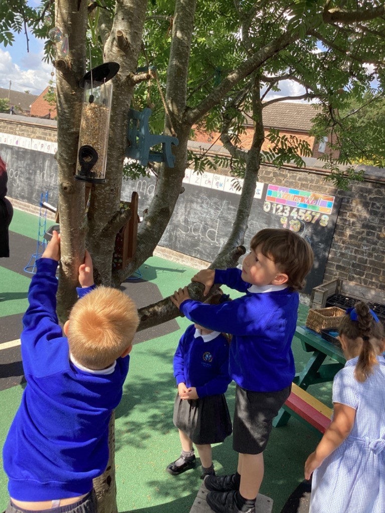 In the image, there are four children outdoors, dressed in blue school uniforms. Two boys are actively engaged with a tree. A bird feeder is hanging from one of its branches. Another boy and a girl are also present in the scene, observing or waiting. The setting appears to be a school playground, with a brick wall and a "Weaving Wall" with numbers visible in the background. The weather seems sunny.