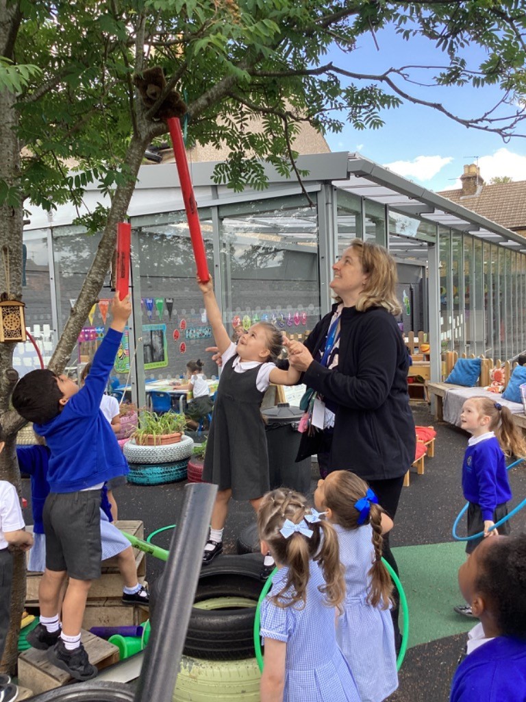 In this image, several children and an adult woman are outdoors, likely in a school or playground setting. One girl is holding a long, red object up towards a tree, where a small, brown, furry object (possibly a teddy bear or similar toy) is positioned in the branches. Another boy is also reaching up with a red object. An adult woman is standing nearby, looking up at the tree and interacting with the girl. Other children are visible in the background and foreground, some holding hula hoops. The setting appears to be an outdoor play area with various equipment, including tires used as planters or play structures, and a building with large windows in the background. The overall impression is one of an engaging and active outdoor learning or play session.