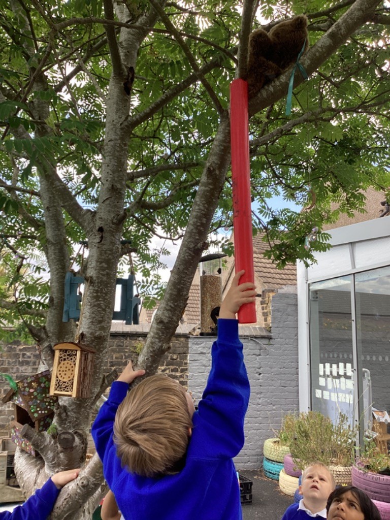In this close-up image, a child is shown reaching up towards a tree branch with a long, red object. Perched on the tree branch above, a brown, fluffy object, possibly a teddy bear or toy, is visible. The child is wearing a blue long-sleeved top. In the background, there's another child looking up, and some outdoor elements like a birdhouse, decorated tires, and a building are faintly visible. The scene appears to be an outdoor activity, likely focused on trying to reach or interact with the object in the tree.