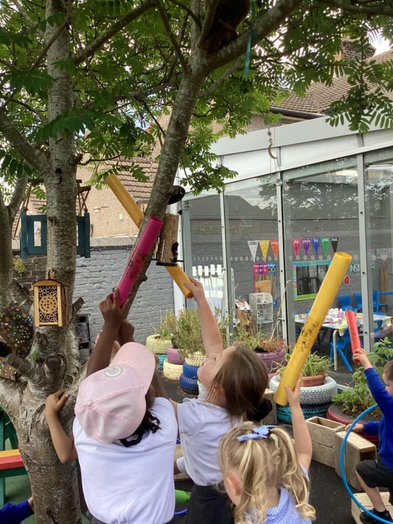 In this image, three children are gathered around a tree, looking upwards and holding various long, cylindrical objects, some of which appear to be pool noodles or similar lightweight tubes. One child, wearing a pink cap, is holding a pink tube upwards. Another child is holding a yellow tube, also directed towards the tree. A third child is in the foreground, with her back to the camera. It looks like they are trying to reach something in the tree, possibly the brown, fluffy object (which appeared in previous images) that can be vaguely seen in the upper branches. The setting is an outdoor play area with painted tires and a building with large windows in the background. There's also a decorative birdhouse hanging from the tree. The children seem engaged in a playful activity.
