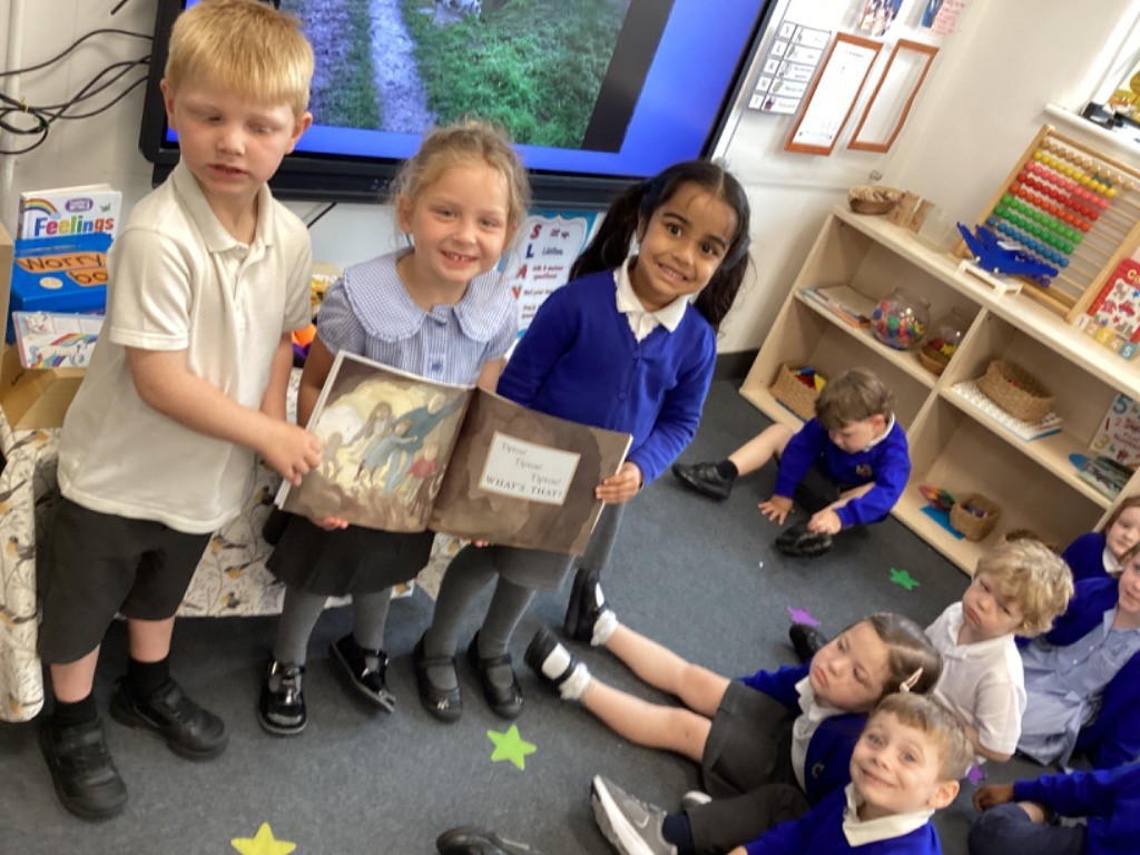 In this indoor classroom setting, several young children are present. In the center, two girls are holding an open book and smiling at the camera. One girl is wearing a light blue and white checkered dress, and the other is in a blue long-sleeved top and dark skirt. A boy in a white polo shirt and dark shorts is standing to their left, looking towards the camera. Other children are visible in the background, some sitting or lying on the floor, and one boy is engrossed in an activity on the right side of the image. The classroom features a large screen, shelves with various educational materials, and a "Feelings box" on the left. The floor is carpeted with star-shaped decorations. The overall atmosphere suggests a lively and engaging learning environment.