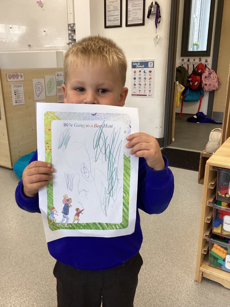In this image, a young girl is holding up a piece of paper with a drawing on it, smiling gently at the camera. The paper has "We're Going on a Bear Hunt" printed at the top, and the girl's drawing includes colorful scribbles and some recognizable figures. She is wearing a blue top. The background shows a classroom setting with wooden tables, chairs, and shelves filled with what appear to be educational materials or toys.