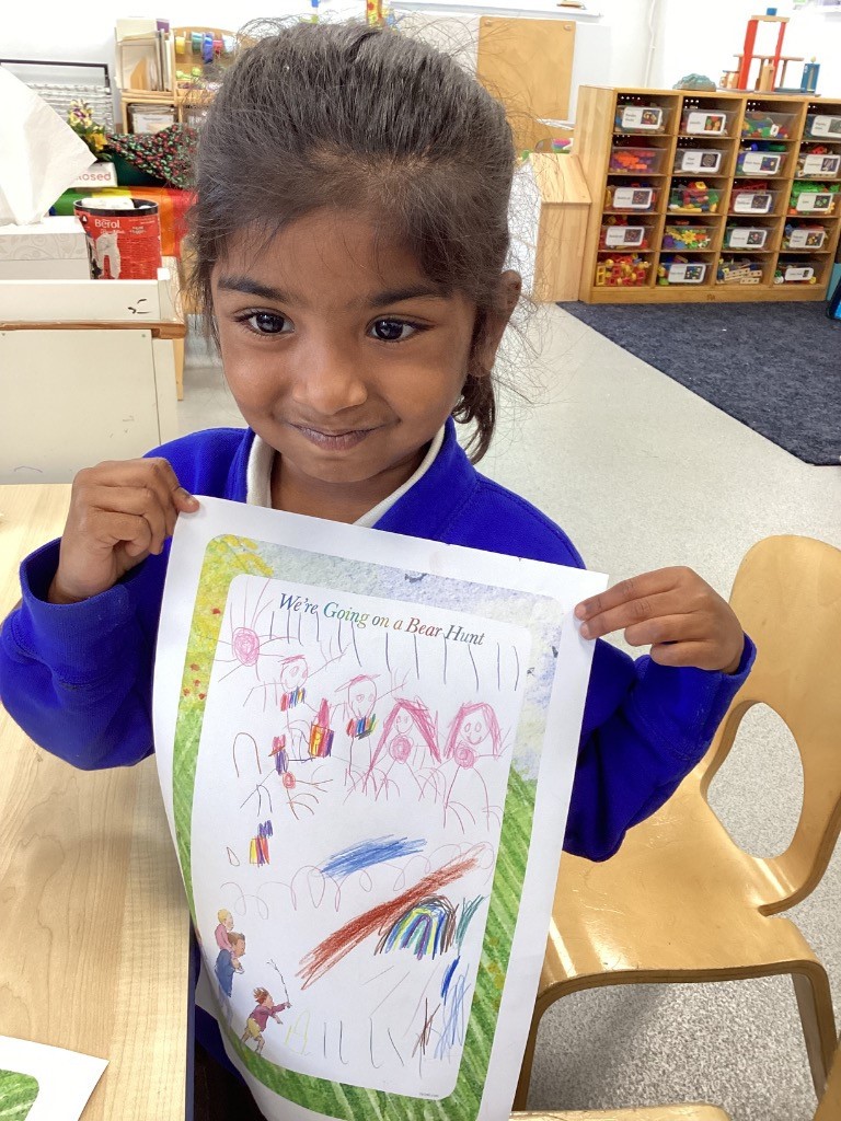 In this image, a young girl is holding up a piece of paper with a drawing on it, smiling gently at the camera. The paper has "We're Going on a Bear Hunt" printed at the top, and the girl's drawing includes colorful scribbles and some recognizable figures. She is wearing a blue top. The background shows a classroom setting with wooden tables, chairs, and shelves filled with what appear to be educational materials or toys.