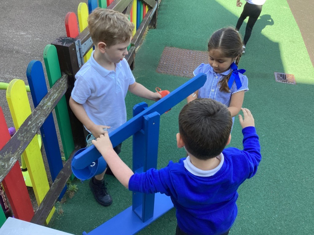 This image shows three young children playing outdoors, likely in a school or playground setting. One boy in a white polo shirt is on the left, looking at a blue seesaw-like play equipment. Another boy in a blue long-sleeved top is in the foreground, facing away from the camera, interacting with the same blue equipment. A girl in a blue and white checkered dress with her hair tied in a ponytail with a blue ribbon is on the right, also engaging with the blue play equipment. The children are on a green surface, possibly artificial turf, and there's a colorful wooden fence behind the boy in the white shirt.
