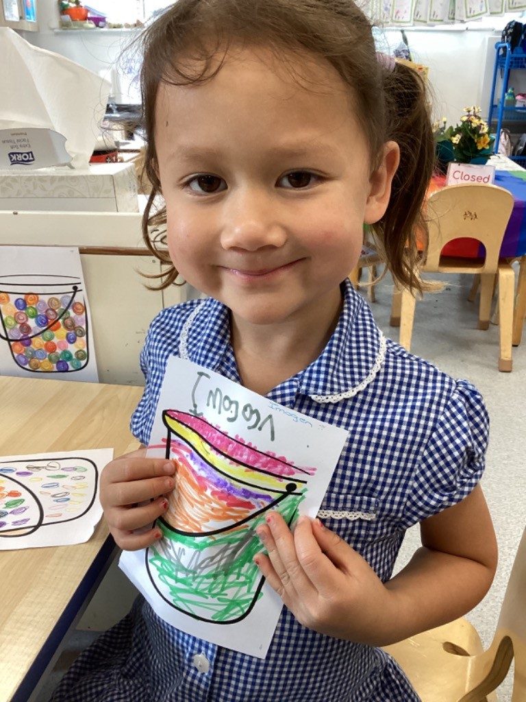 This image shows a young girl holding up a colorful drawing of a bucket. The girl is smiling and looking at the camera. She is wearing a blue and white checkered uniform dress. The drawing she's holding is a cheerful depiction of a bucket filled with various colored items, and the word "Viggo" is written on it. Another similar drawing is visible on the table to her left. The setting appears to be a classroom, with a table, chairs, and various items in the background, including a "Closed" sign on a colorful chair.