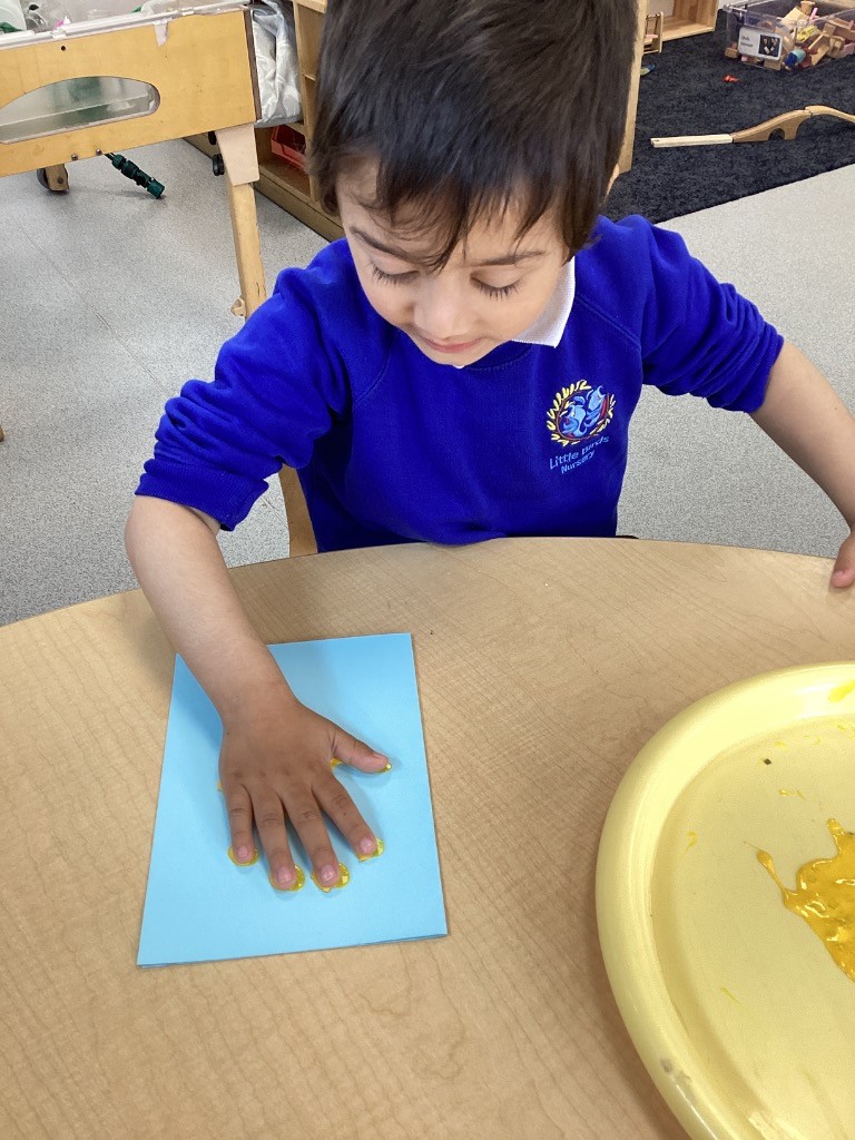 This image shows a child in a blue school uniform, with a white collar and a school emblem, engaged in an art activity at a light-colored wooden table. The child is pressing their hand onto a blue sheet of paper, with yellow paint visible on their fingertips and the paper, suggesting they are making a handprint. To the right, there is a yellow tray with some spilled yellow paint. The background shows a classroom setting with wooden furniture and other learning materials.