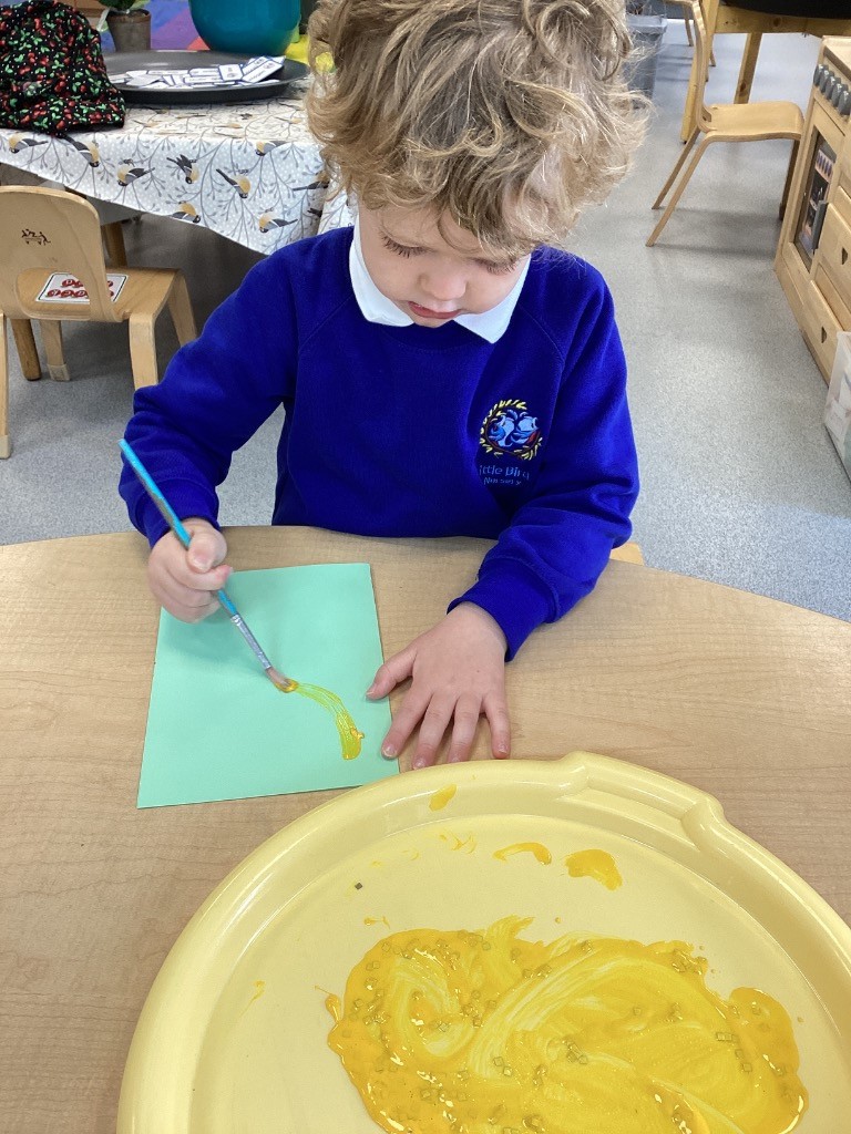 This image shows a child in a blue school uniform with a white collar and a school emblem, seated at a light-colored wooden table and engaged in painting. The child is holding a paintbrush and applying yellow paint to a light green sheet of paper. On the table, there's a yellow tray with a pool of yellow paint. The background suggests a classroom environment, with other tables and chairs visible.