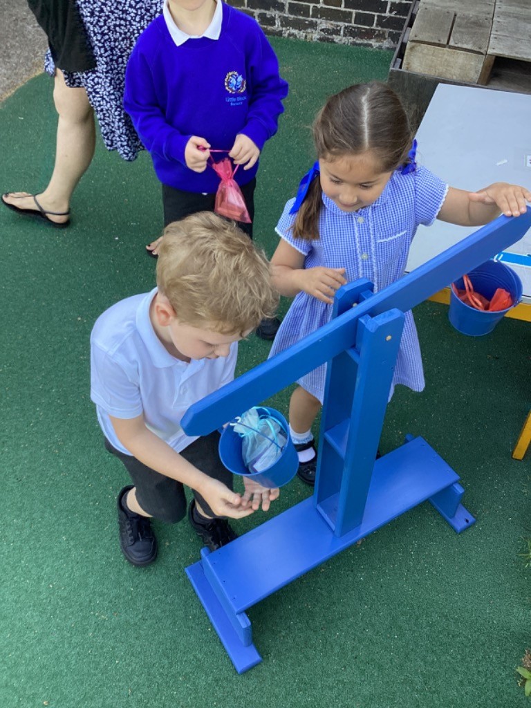 This image features two children and an adult's legs in the background, interacting with a blue balance scale-like play equipment outdoors. A boy in a white polo shirt is kneeling on the left, placing something into a blue bucket attached to one end of the balance scale. A girl in a blue and white checkered dress, with a blue ribbon in her hair, is on the right, also engaging with the equipment. Another child in a blue uniform and an adult are partially visible in the background on the left. The setting appears to be an outdoor play area with a green artificial turf surface.
