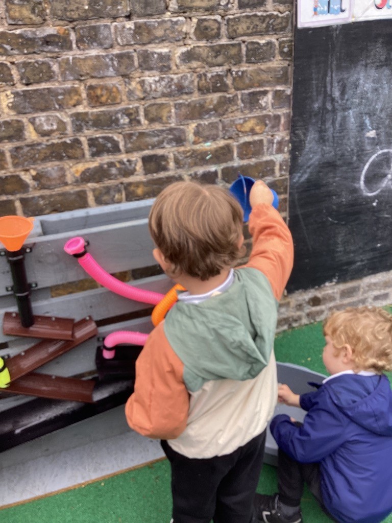 This image shows two young children playing with a water or sensory wall outdoors. One child, seen from behind, is wearing an orange and light green hooded jacket and is holding a blue cup, appearing to pour something into the water wall structure that has tubes, funnels, and channels. Another child, with curly blonde hair, is partially visible on the right, kneeling and engaging with a grey container on the ground. The background features a brick wall on the left and a dark surface, possibly a chalkboard, on the right. The ground is covered with green artificial turf.