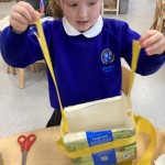 This image shows a child in a blue school uniform, with a white collar and a school emblem, holding up a craft project. The child appears to be making a bag or container out of what looks like a repurposed milk carton or similar rectangular box, which is wrapped with yellow ribbon. The child is holding the yellow ribbon straps as if to form a handle. On the table, there are red-handled scissors. The background suggests a classroom or learning environment.