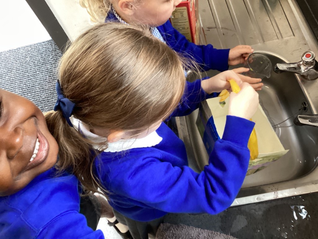 This image shows two children in blue school uniforms at a sink, engaged in an activity that involves water. One child, seen from the back, has light brown hair tied in a ponytail with a dark blue bow and is wearing a blue top with a white collar. They are holding a yellow object and what looks like a repurposed carton or box under the running water from a tap in a metal sink. Another child with lighter, curly hair is partially visible behind the first, also interacting with the sink. A third child is partially visible on the far left, looking towards the camera and smiling. The setting appears to be a classroom or utility area within a school.