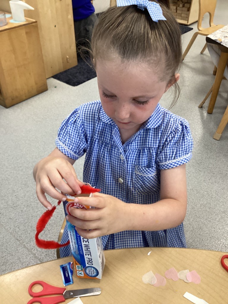 This image shows a child in a blue and white checkered uniform dress, with a blue bow in their hair, engaged in a crafting activity at a wooden table. The child is focused on decorating a small carton, possibly a juice box or similar container, with red pipe cleaners. On the table, there are also a pair of red-handled scissors, some small white and pink heart-shaped cutouts, and other crafting bits. The setting appears to be a classroom, with furniture and possibly other learning materials in the background.