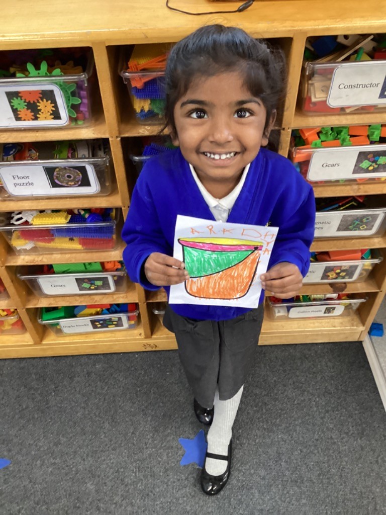 This image shows a smiling child holding up a drawing of a bucket. The child is wearing a blue top, white shirt, and grey trousers with white socks and black shoes. They are standing in front of a wooden shelving unit filled with clear plastic containers holding various toys and learning materials, some of which are labeled "Constructor," "Gears," and "Floor puzzle." The drawing held by the child is colorful and depicts a bucket with different colored sections.