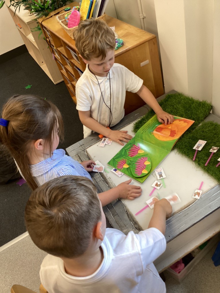 two children are looking at an open book with illustrations, and there are small picture cards on a grassy mat next to the book, possibly for sequencing the story.