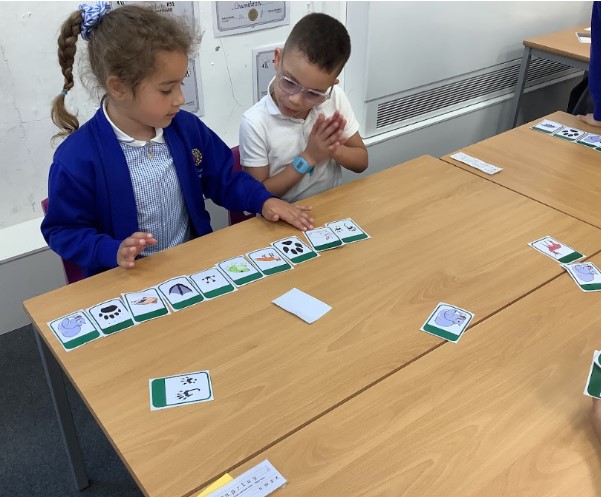 This image shows two young students at a wooden table in a classroom, engaged in what appears to be a matching or sequencing game. A student on the left, wearing a blue school uniform, is reaching out towards a line of cards laid out on the table. These cards feature images of animals and their footprints. The student on the right, wearing a white shirt and glasses, is looking down at the cards with hands clasped together, seemingly observing or thinking. There are more cards scattered on the table, some of which appear to be face down or in different arrangements. The background shows a classroom wall with some papers or certificates.