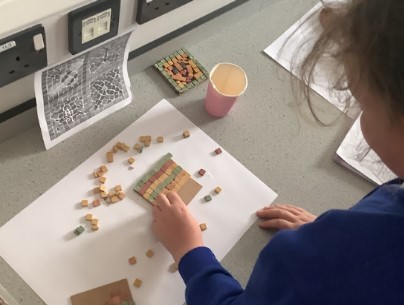 This image shows a child, seen from behind, engaged in a creative activity at a table. The child is wearing a blue top and appears to be working with small, square-shaped pieces, possibly made of wood or mosaic tiles, on a piece of cardboard. They are creating a pattern or design. On the table, there are loose square pieces, a pink paper cup, and another finished or in-progress mosaic piece. In the background, on the wall, there's a black and white patterned print, possibly a reference or example for the activity, and electrical outlets. The scene suggests a craft or art lesson in a classroom setting.