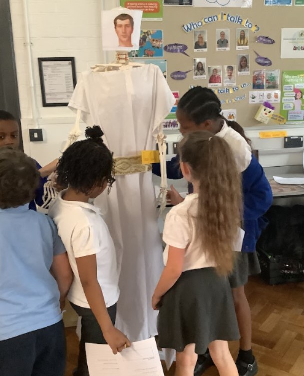 This image shows a group of children gathered around a life-sized skeleton model dressed in a white robe, possibly resembling a toga, and a golden belt. A printed image of a human head is attached to the top of the skeleton, giving it the appearance of a historical figure. The children are observing and interacting with the display, suggesting a learning activity, possibly related to ancient history, anatomy, or a cultural theme. One child on the left is partially obscured, while others are closer to the display, looking at it intently. The setting is a classroom, with posters and displays on the wall in the background.