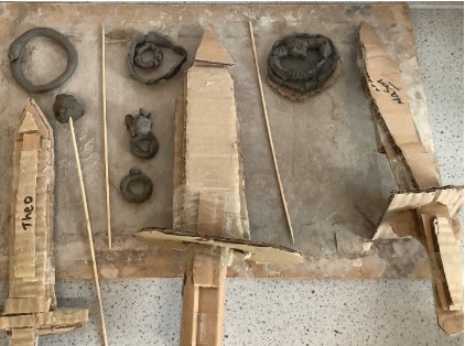 This image displays a collection of handmade objects on a wooden surface, suggesting a classroom art or craft project. The objects include: Several items made from what appears to be clay or playdough, including rings, amorphous shapes, and possibly a small bowl or coil pot. Multiple swords crafted from cardboard. One sword on the left has "Theo" written on it. Long, thin wooden sticks, possibly skewers or dowels, placed among the objects. The overall impression is that these are creations from a school project, perhaps related to a historical theme (like ancient tools or weapons) or simply a creative art activity.