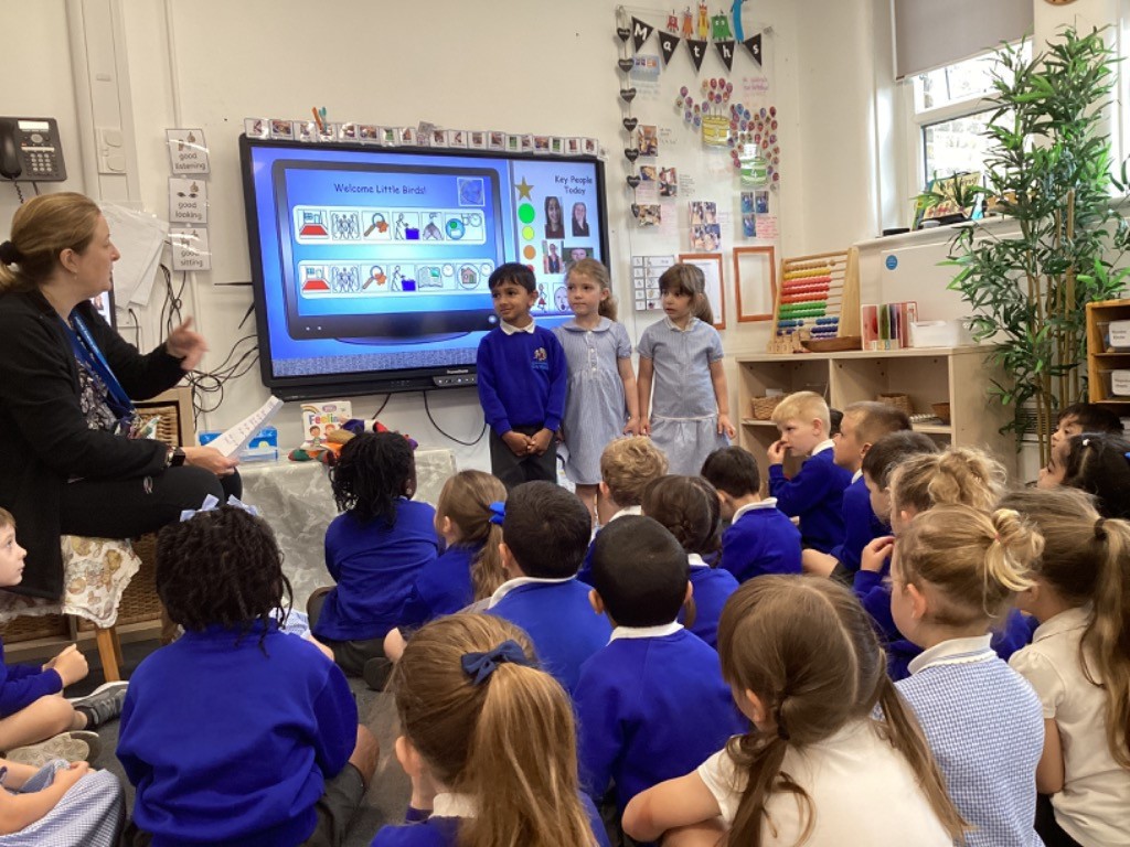 The image captures a vibrant classroom scene, likely in an early years or primary school. A teacher, seated on the left, is addressing a group of young children who are sitting on the floor, attentively looking towards the front of the room. In front of a large interactive screen, three children are standing. The screen displays "Welcome Little Birds!" and "Key People Today," along with various icons and images, suggesting an interactive lesson or a daily routine presentation. The classroom is brightly lit, with natural light coming from a window on the right. There are also educational resources visible, such as an abacus and shelves filled with learning materials. The overall atmosphere appears to be one of engaging and structured learning.