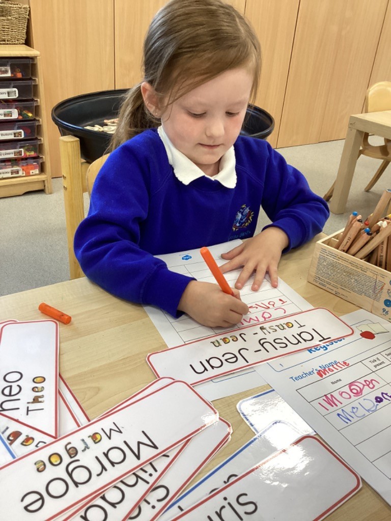 A young girl is sitting at a desk, writing on a piece of paper that has names printed on it, including "Tansy-Jean" and "Margoe." She might be practicing writing or taking attendance.