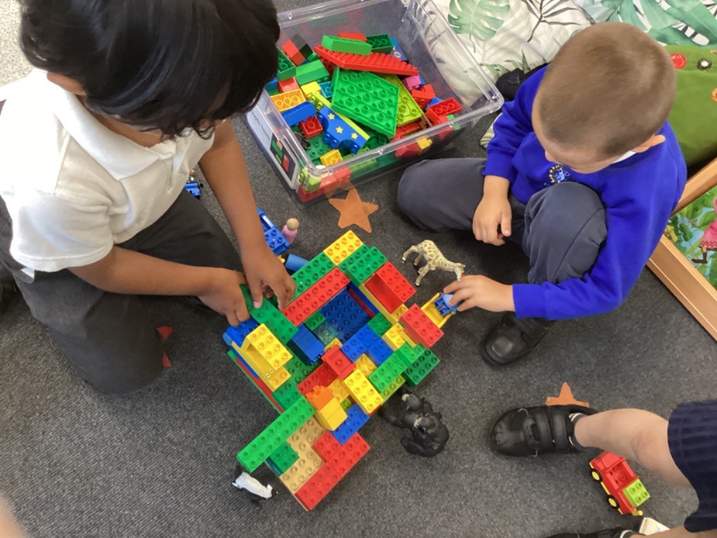 In the image, two young boys are sitting on a carpeted floor, engrossed in playing with a large pile of colorful building blocks, likely LEGO or Duplo. They have a clear plastic container filled with more blocks next to them. One boy, on the left, is wearing a white polo shirt and is actively building with the blocks. The other boy, on the right, is wearing a blue long-sleeved top and dark trousers, and is also engaged in building, with some animal figures also visible among the blocks.