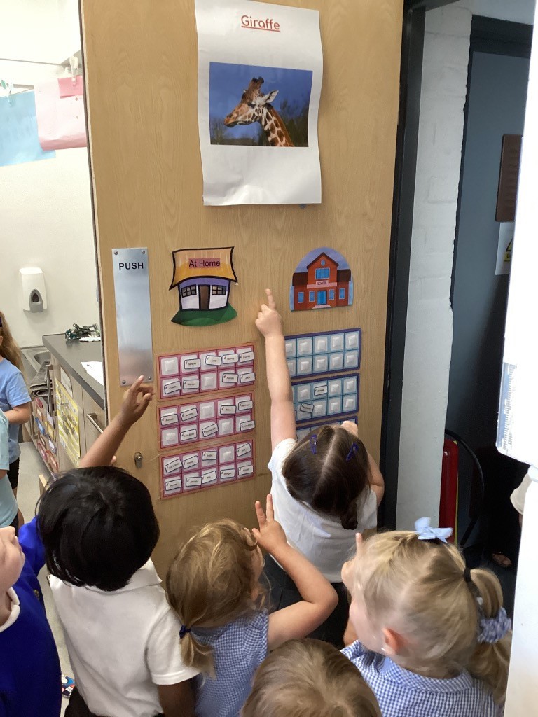 A group of children are gathered in front of a door with a picture of a giraffe and some charts below it. They are pointing at the charts, suggesting some kind of interactive learning or tracking activity.