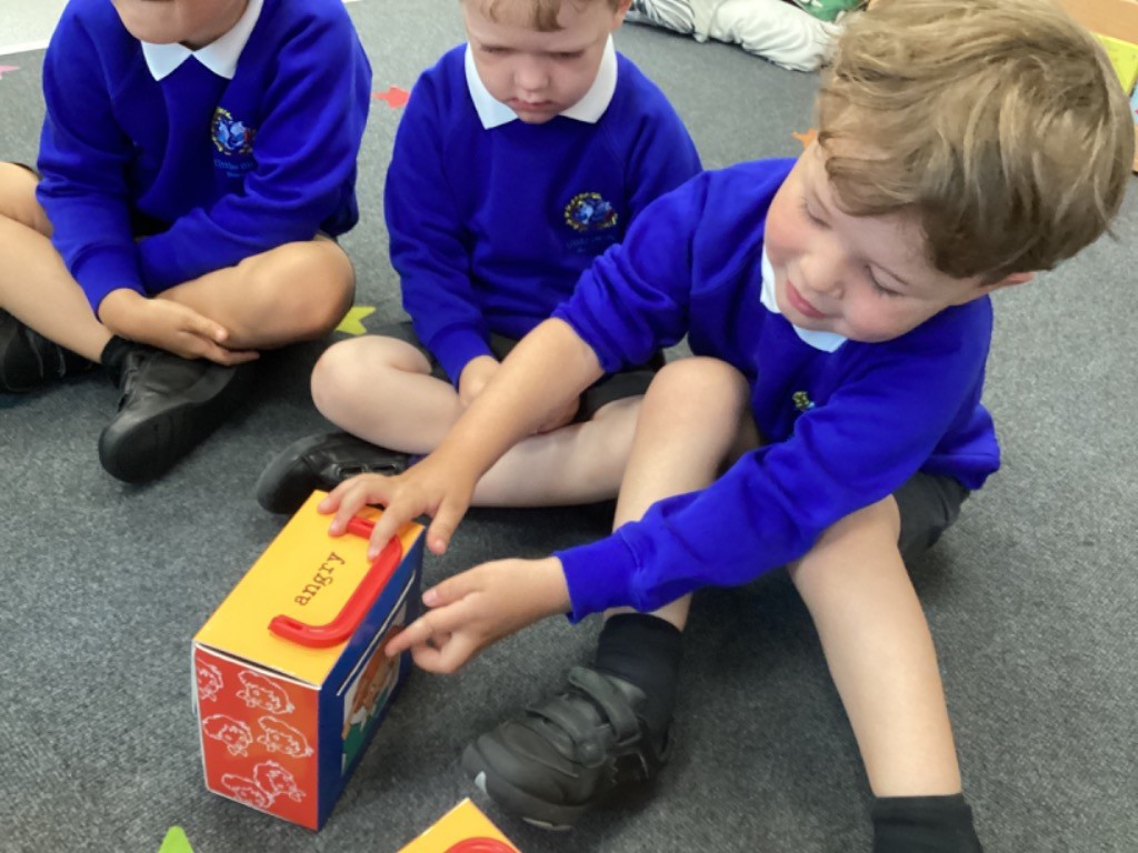 Three young boys are sitting on the floor. One boy in the foreground is interacting with a colorful box that has the word "angry" on it, possibly a tool for understanding emotions.