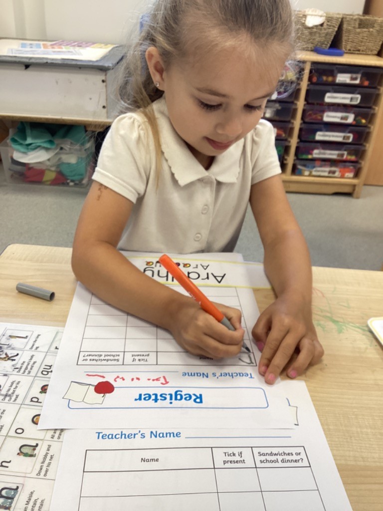 A young girl is at a desk, using a marker to fill out a "Register" sheet that includes spaces for "Teacher's Name" and a tick box for "Sandwiches or school dinner?". She is likely practicing taking attendance or learning about daily routines.