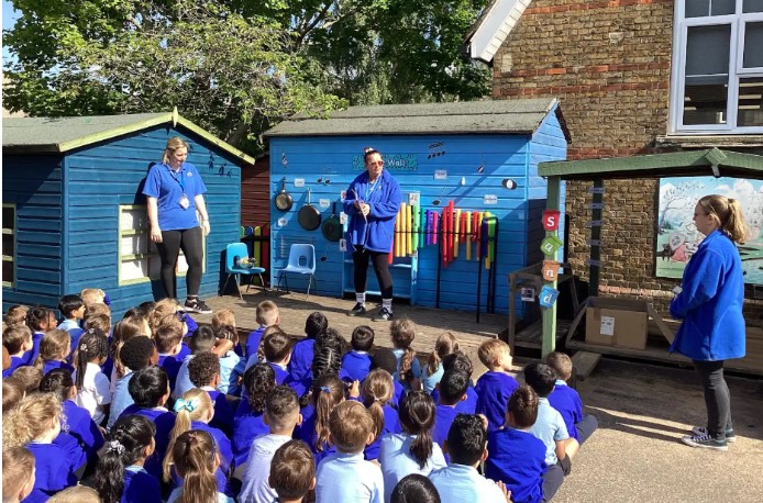 This image depicts an outdoor educational or entertainment activity, likely at a school. A large group of young children, predominantly wearing blue tops, are sitting on the ground, facing a small raised wooden stage or platform. On the platform, two adults are present. One adult, wearing a blue t-shirt and black trousers, is standing on the left side of the platform. The other adult, also in a blue top, is standing more centrally and appears to be leading the activity, possibly interacting with the children. Behind the stage, there are two blue wooden sheds. The shed on the right has various musical instruments or sound-making objects attached to its exterior, such as pans, cymbals, and colorful tubes, suggesting it might be a "sound wall" or similar interactive display. To the far right, another adult, dressed in a blue jacket, is standing near a wall or building. The setting appears to be a school playground, with brick buildings visible in the background and trees overhead.