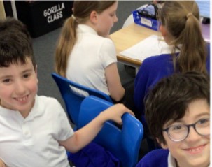 This image shows several children in a classroom setting, likely during school. In the foreground, two boys are looking towards the camera and smiling. The boy on the left has dark curly hair and is wearing a white polo shirt. The boy on the right wears glasses and also a white polo shirt. In the background, other students are visible at a table, appearing to be engaged in an activity or conversation. One girl has her back partially to the camera, wearing a white top. Another student, possibly wearing a blue uniform, is also visible.