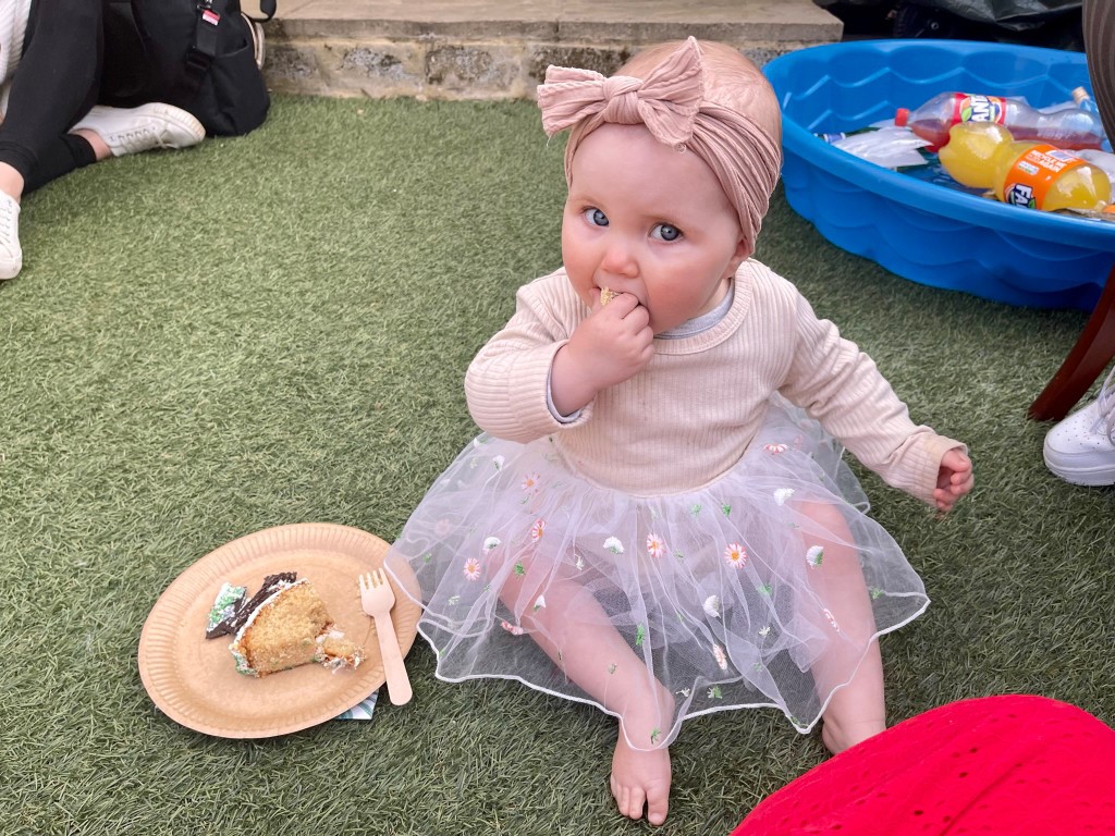 A baby is sitting on green artificial grass, eating from their hand. In front of the baby is a paper plate with a slice of cake. The baby is wearing a light-colored top and a white tutu skirt with small daisy designs, and a pink bow on their head. A small blue paddling pool and some drinks are visible in the background.