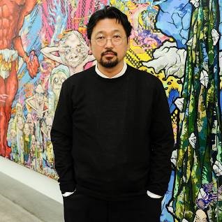 Artist Takashi Murakami. He is a man with a beard and glasses, wearing a black long-sleeved shirt, is standing in front of a colorful, abstract painting.