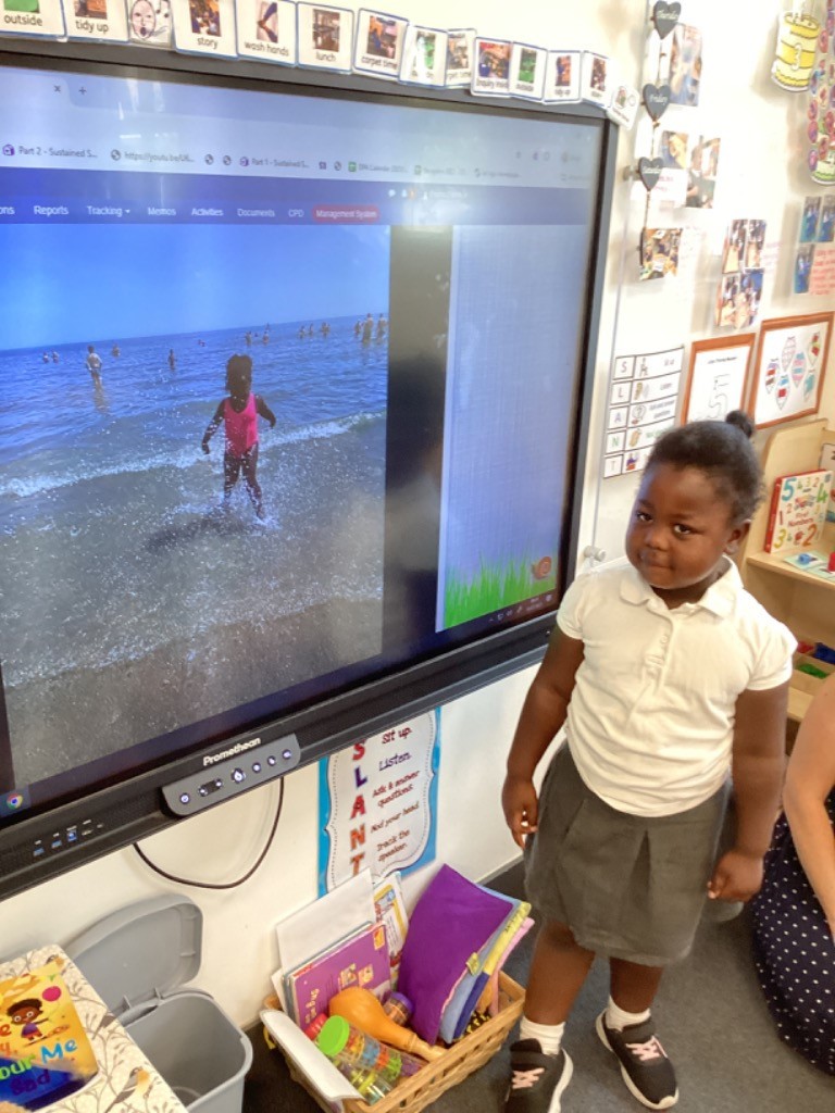 This image shows a child in a white polo shirt and grey skirt standing next to a large interactive screen displaying an image of a child in the sea. This suggests a learning activity involving visual media, perhaps discussing a place, an experience, or a story. There are also classroom decorations and learning aids visible on the wall.