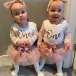 Two babies, possibly twins, are standing side-by-side. Both are wearing matching long-sleeved tops with "One" written on them, pink tutus, and pink bows in their hair, suggesting a first birthday celebration.