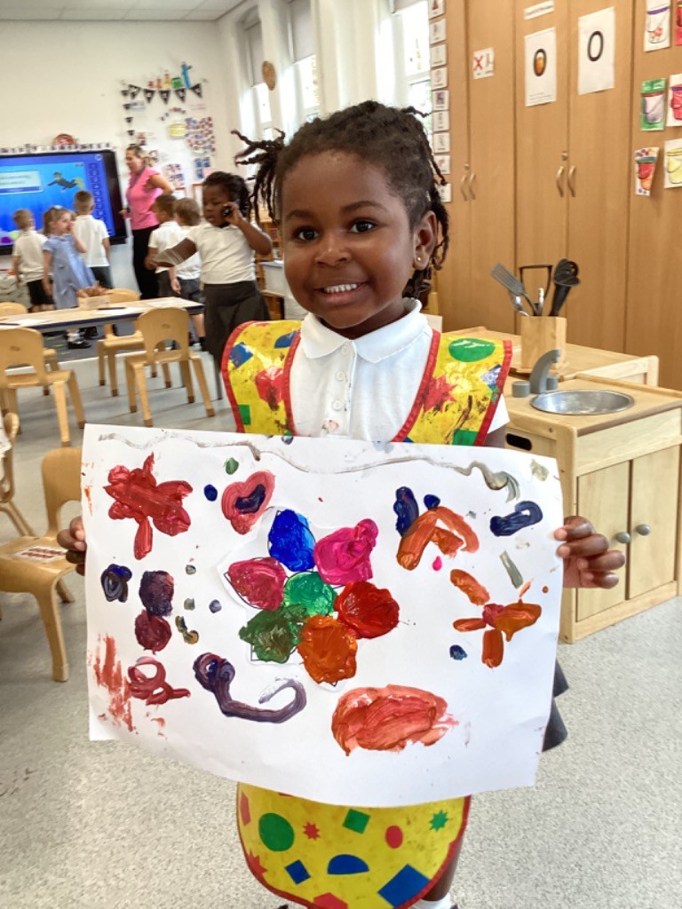 A young girl, wearing a white collared shirt and a yellow art smock covered in paint, is standing and proudly holding up a white sheet of paper with a colorful painting on it. She is smiling at the camera.