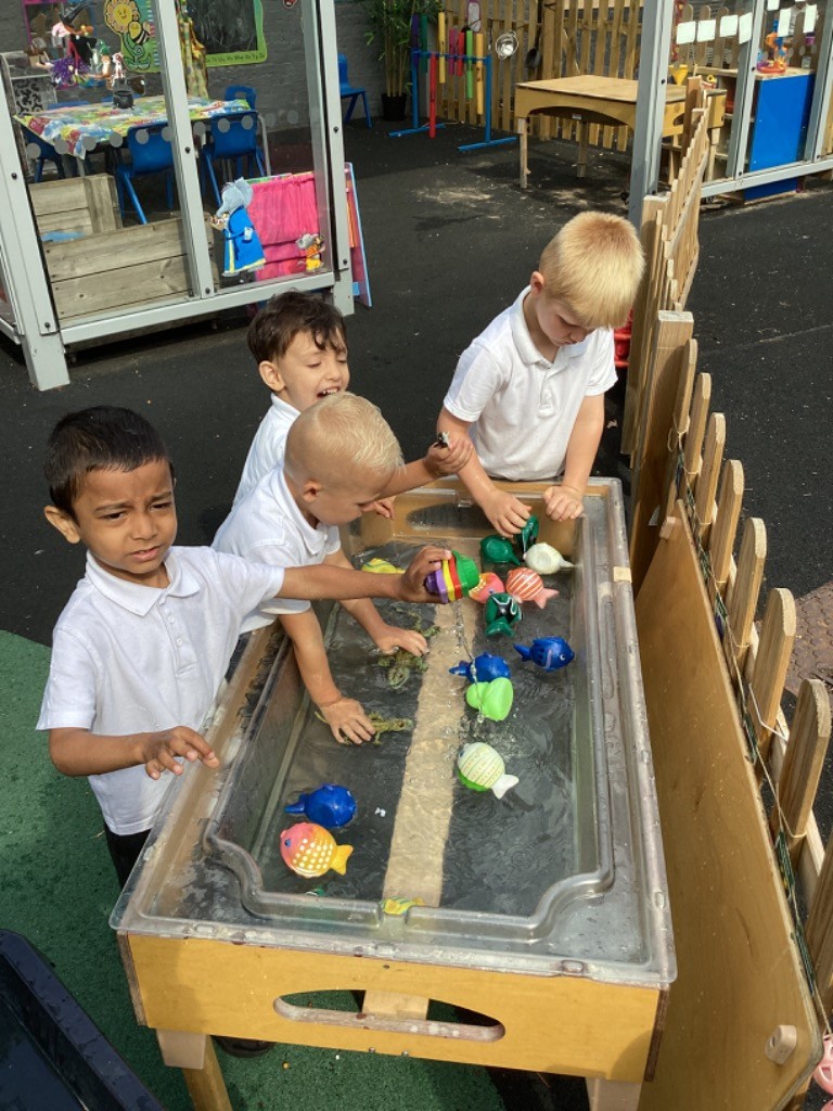 This image shows three boys, dressed in white polo shirts, playing at a water table outdoors. They are manipulating various colorful plastic toy fish and other objects in the water, indicating sensory play and exploration of water properties.