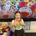 A young girl with pigtails is smiling and holding a colorful paper flower craft. She is standing in front of a large screen displaying vibrant, stylized artwork with smiling flowers and cartoon characters.