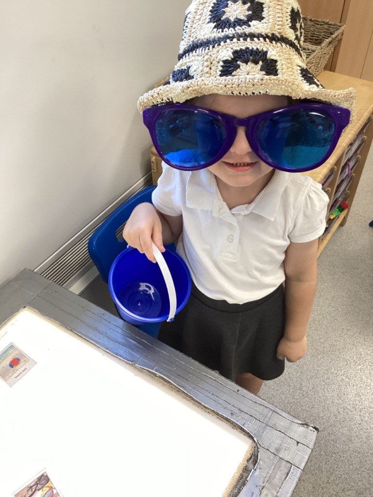 This image features a young child wearing a white polo shirt and a dark skirt, topped with a large, crocheted straw hat and oversized blue sunglasses that cover most of their face. The child is holding a blue bucket and stands next to a light table. The expression visible suggests playfulness.
