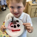 A young boy with light brown hair and blue eyes, wearing a white polo shirt, is holding up a paper plate with a crafted face on it, smiling at the camera. The craft seems to involve paint and some fuzzy material for hair.
