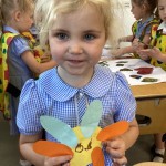 A young girl with blonde hair, wearing a blue and white checkered dress, is smiling at the camera while holding a colorful paper craft that looks like a whimsical creature with multi-colored petals or wings.