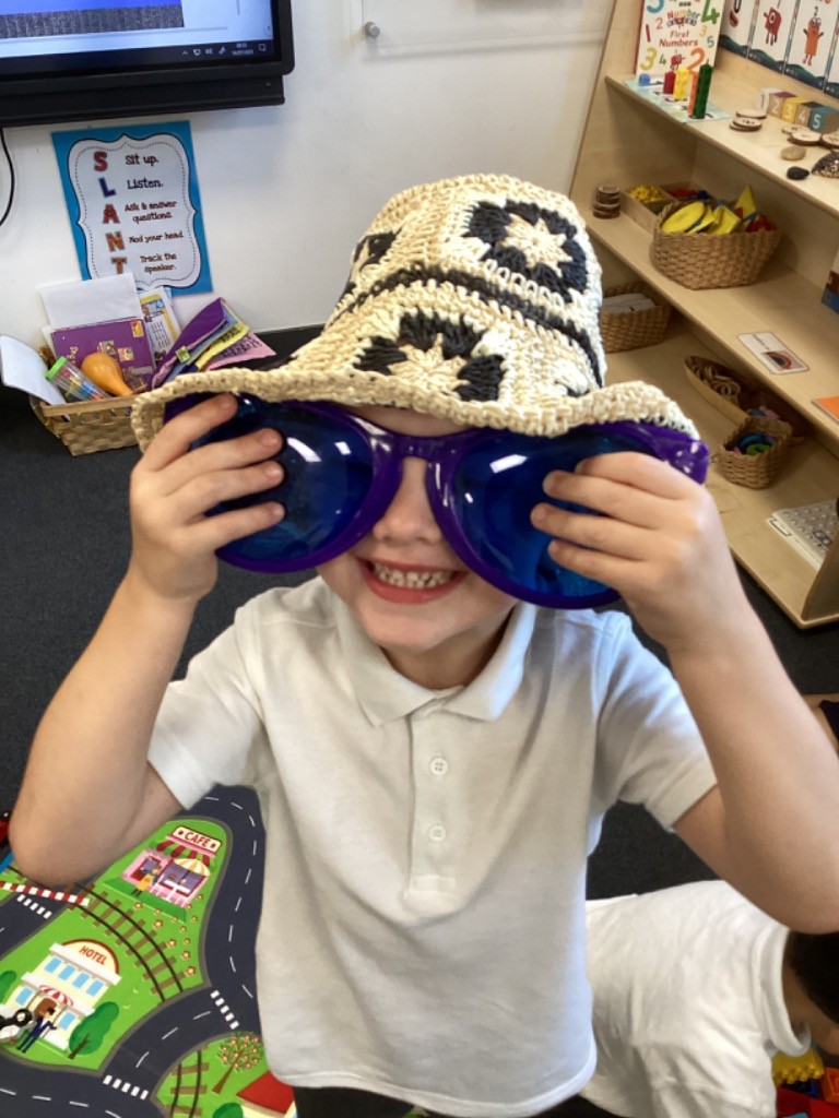 This is a close-up of the a child wearing the crocheted hat and the large blue sunglasses. The child is smiling widely, holding the sunglasses up to their face, further emphasizing the fun and playful nature of the activity. Part of a playmat with roads and buildings is visible on the floor below.