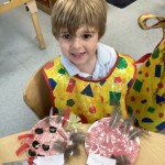 A young boy with light brown hair, wearing a colorful art smock, is sitting at a table with two paper plate crafts in front of him, looking up and smiling at the camera. The crafts appear to have paint and feathers.
