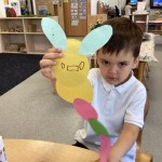 A young boy, wearing a white polo shirt, is holding up a craft project that resembles a yellow caterpillar with light blue ears/antennae and a long, colorful body made of paper circles. He is looking directly at the camera with a serious expression.