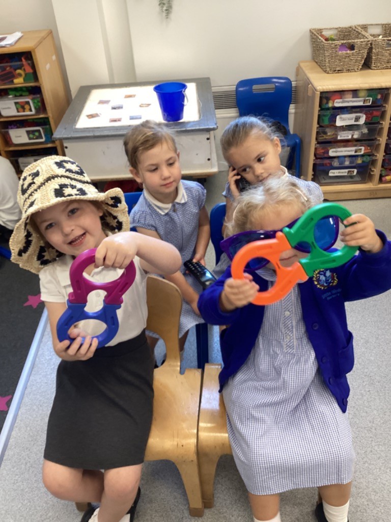 This image shows a group of four children holding two brightly colored, oversized magnifiers (or possibly magnetic toys) in purple and blue. To their right, another child in a blue and white checkered dress is seated. On the far right, a child in a blue uniform jacket is also holding two large magnifiers (orange and green). Behind them, another child is seated and appears to be pretending to talk on a phone. The children are clearly engaged in imaginative play, possibly exploring different perspectives with the magnifiers or simply enjoying dressing up and interacting with oversized props. A light table is also visible in the background.