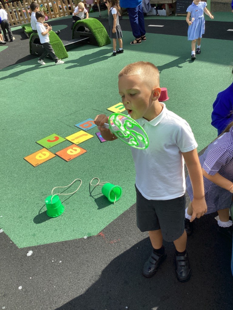 A boy in a white polo shirt and dark shorts is outside on a green playground surface, blowing bubbles with a large, green bubble wand. In the background, there are colorful numbered mats on the ground.